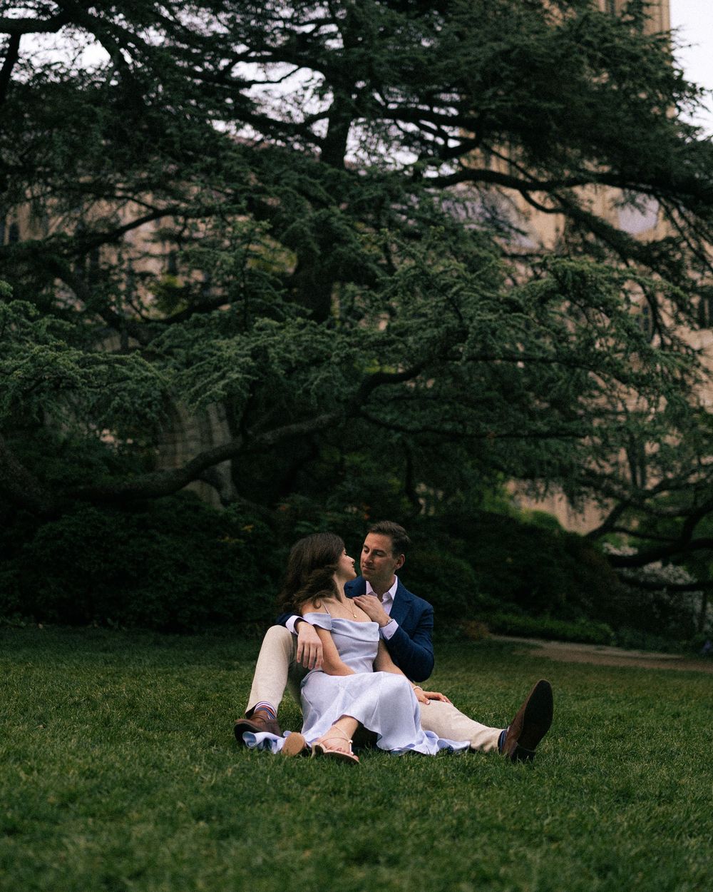 Couple sitting on lush green grass sharing a romantic moment beneath a large cedar tree near a stone building.