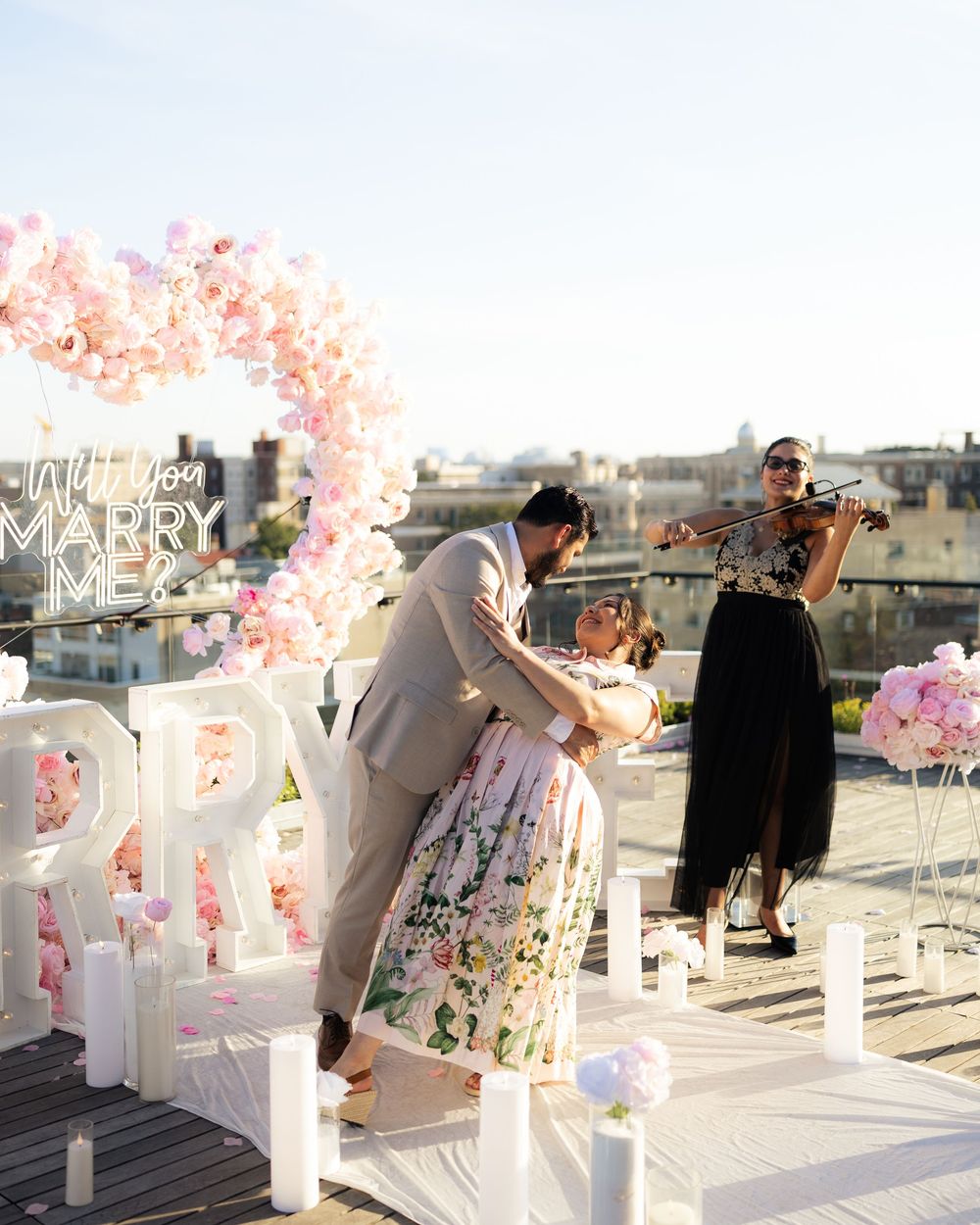 Romantic rooftop proposal with floral arch, neon sign, violinist, and couple sharing a kiss.