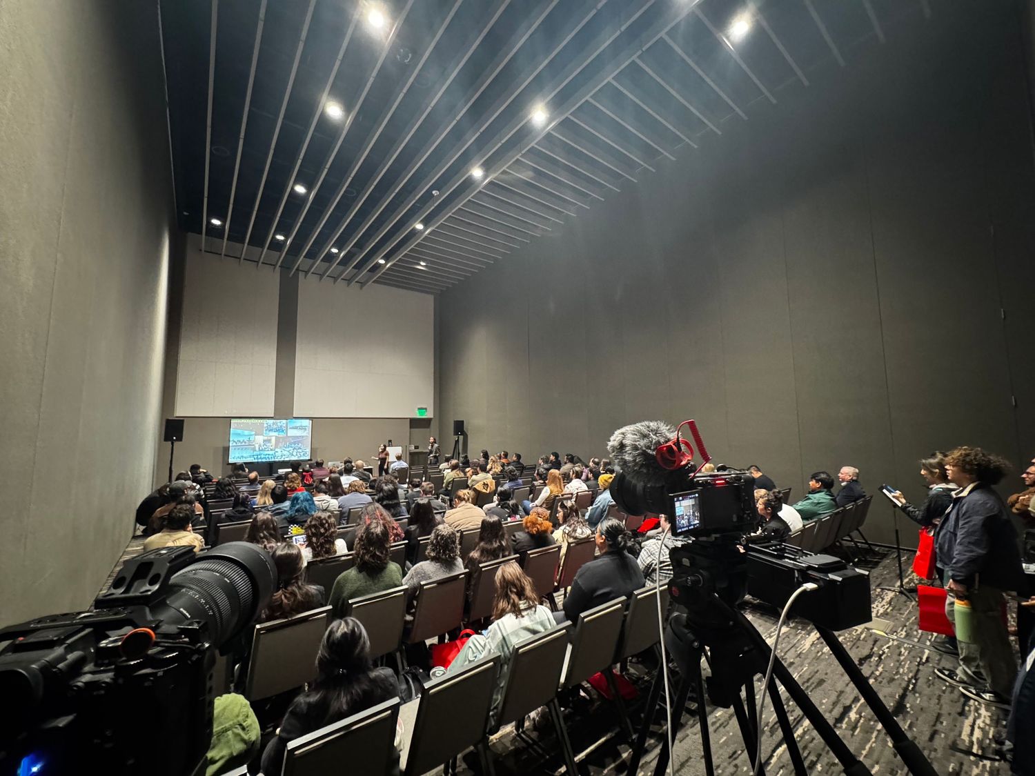 A dark auditorium filled with seated people watching a presentation or lecture with overhead lighting.