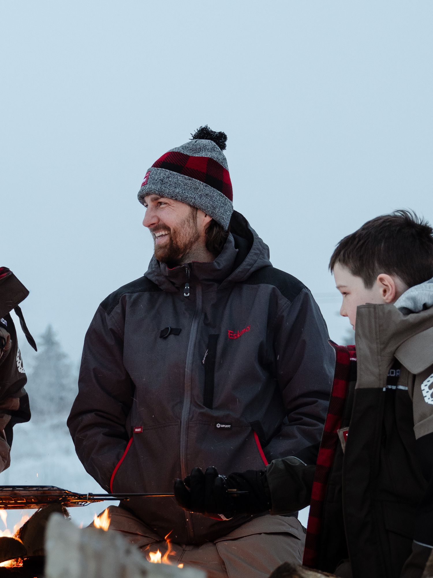 Winter ice fishing scene with people gathered around campfire in snowy conditions.