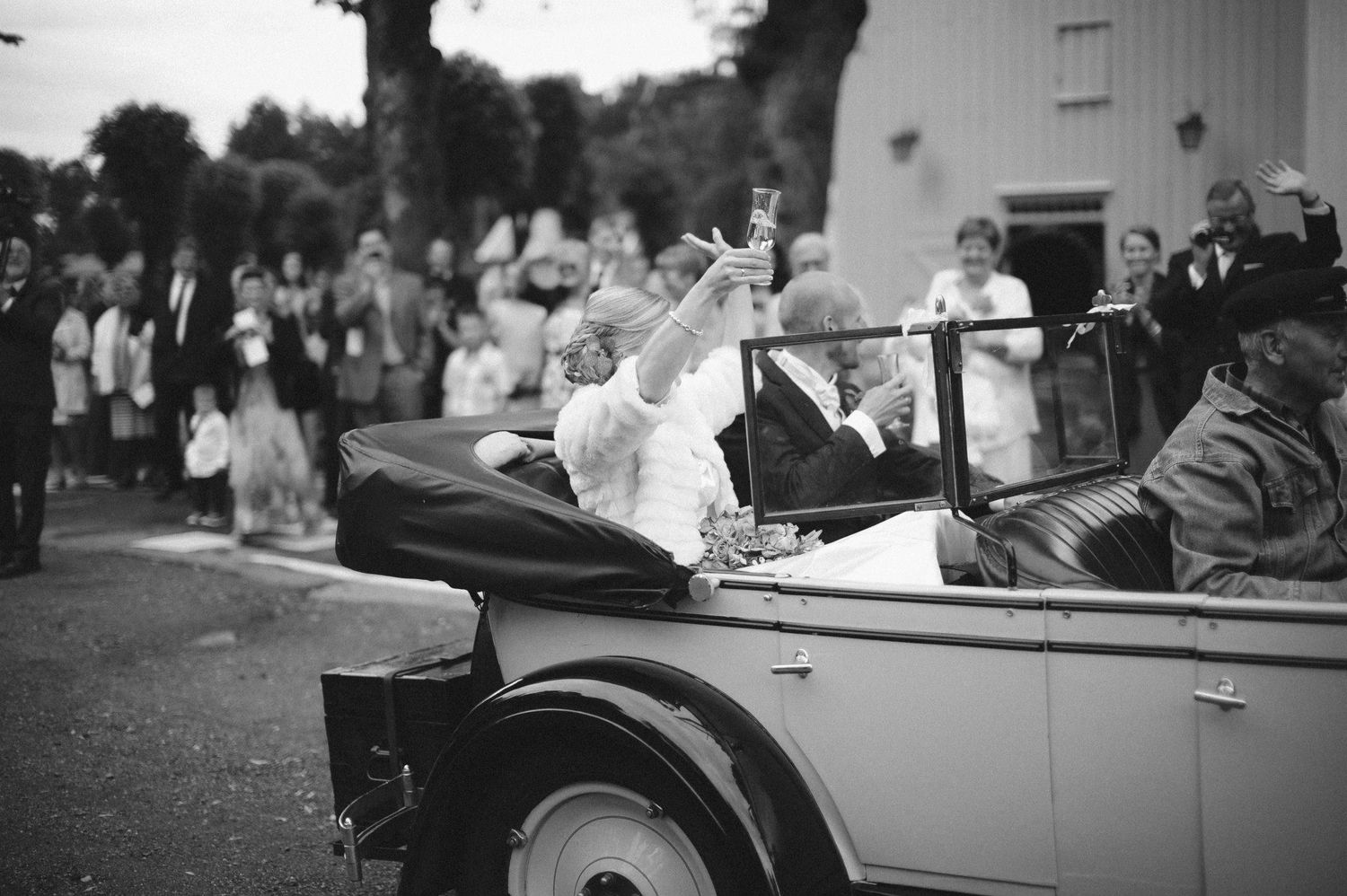 Black and white photo of someone waving from a vintage car during a celebration.