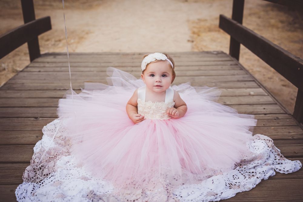 A small figure in a flowing pink tulle and lace dress with white headband sits on wooden bridge.