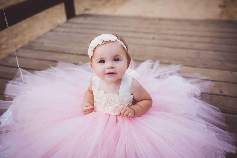 A baby in a pink tulle dress sits on wooden decking while being photographed in multiple poses.