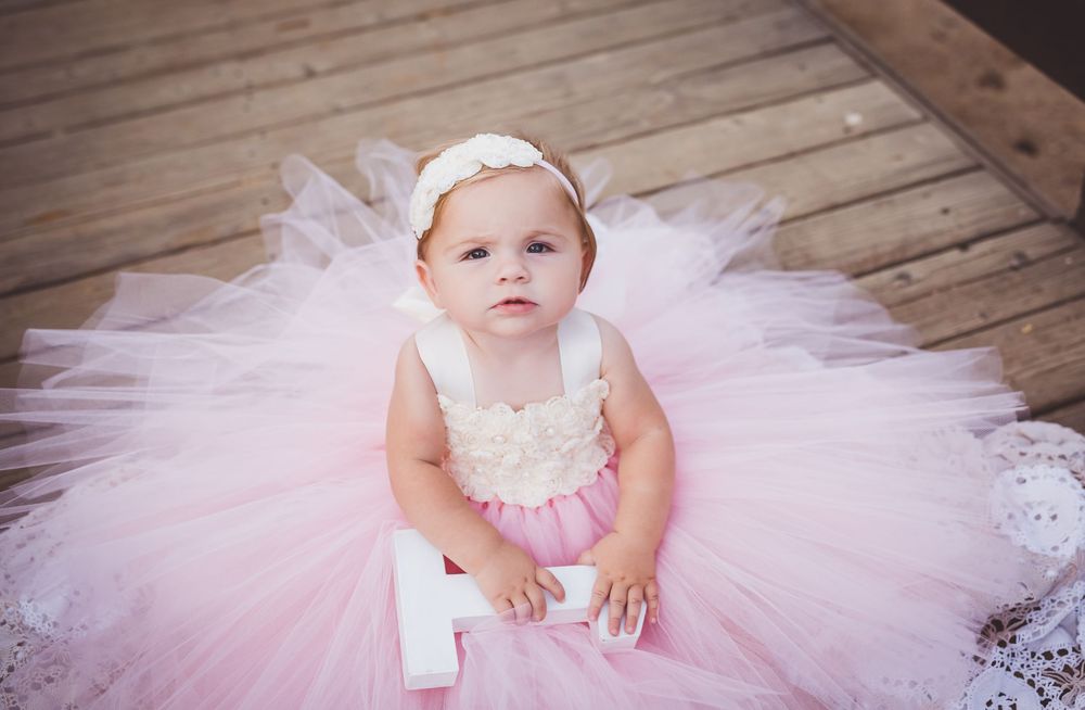 A baby in a pink tutu dress sits on a wooden floor while being photographed in a series of poses.