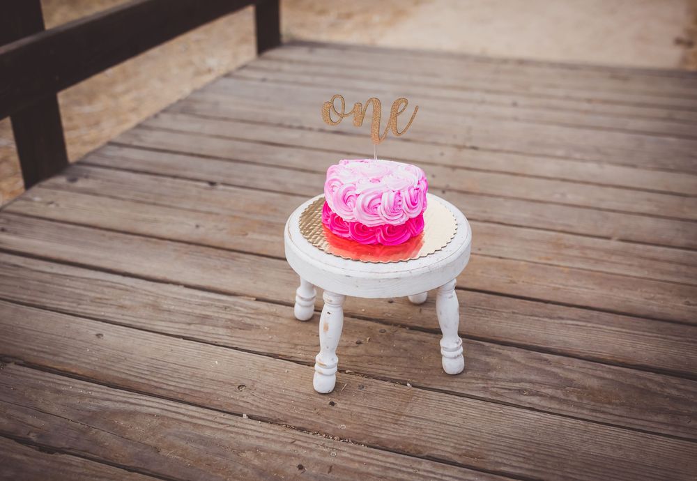 A pink birthday cake with sprinkles sits on a small white decorative stool on a wooden deck.