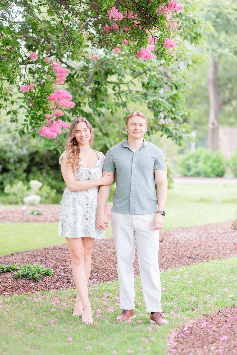 A couple poses together under blooming pink crepe myrtle trees in a garden setting.