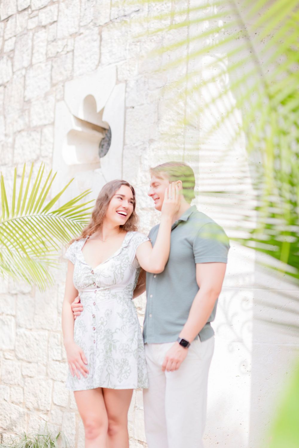 A romantic photoshoot against a white stone wall with palm fronds creating natural framing.