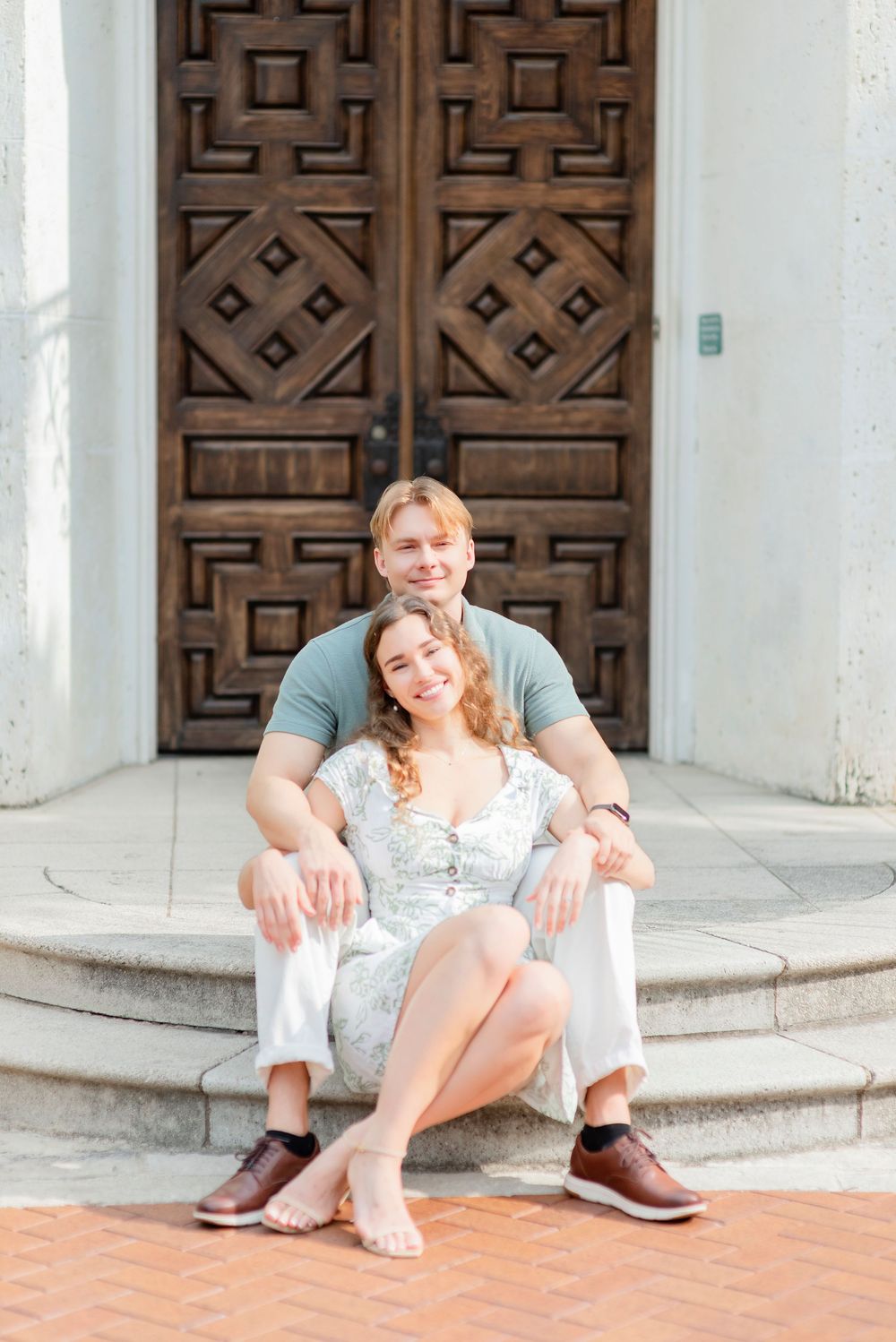A couple sits casually on stone steps in front of ornate wooden doors, creating a relaxed outdoor portrait scene.