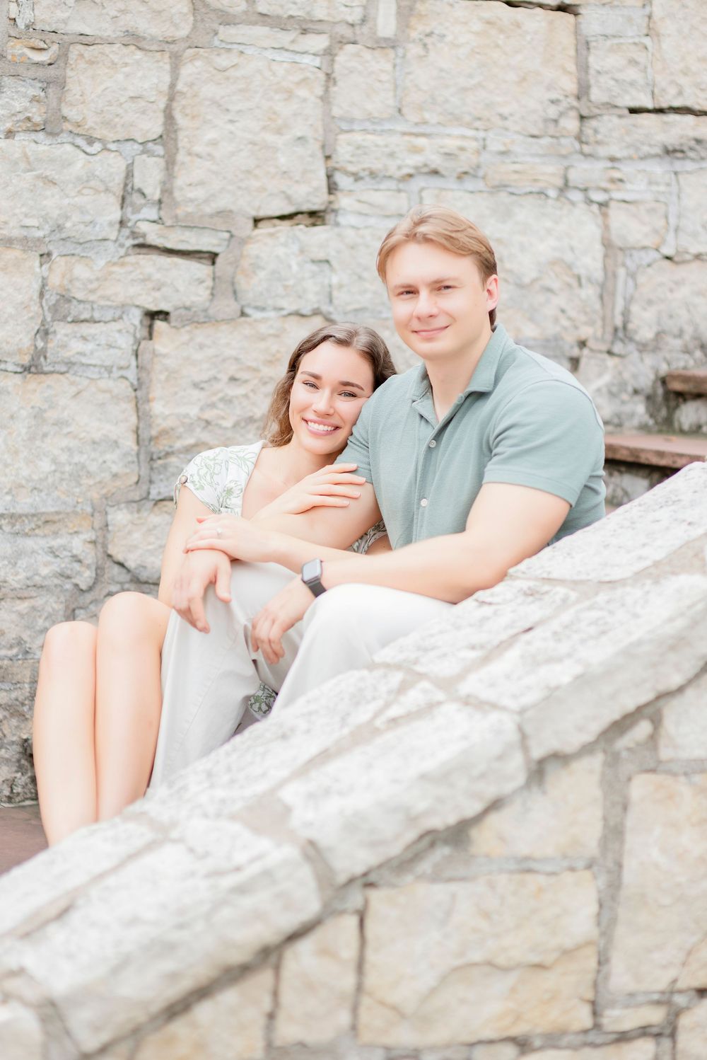 A couple shares romantic moments while sitting on stone steps against a rustic wall background.