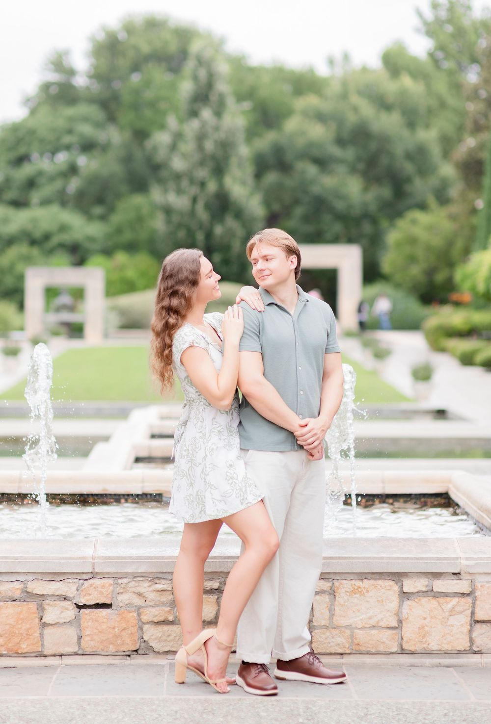 A romantic couple embraces by a stone fountain in a lush garden setting during a sunny afternoon.