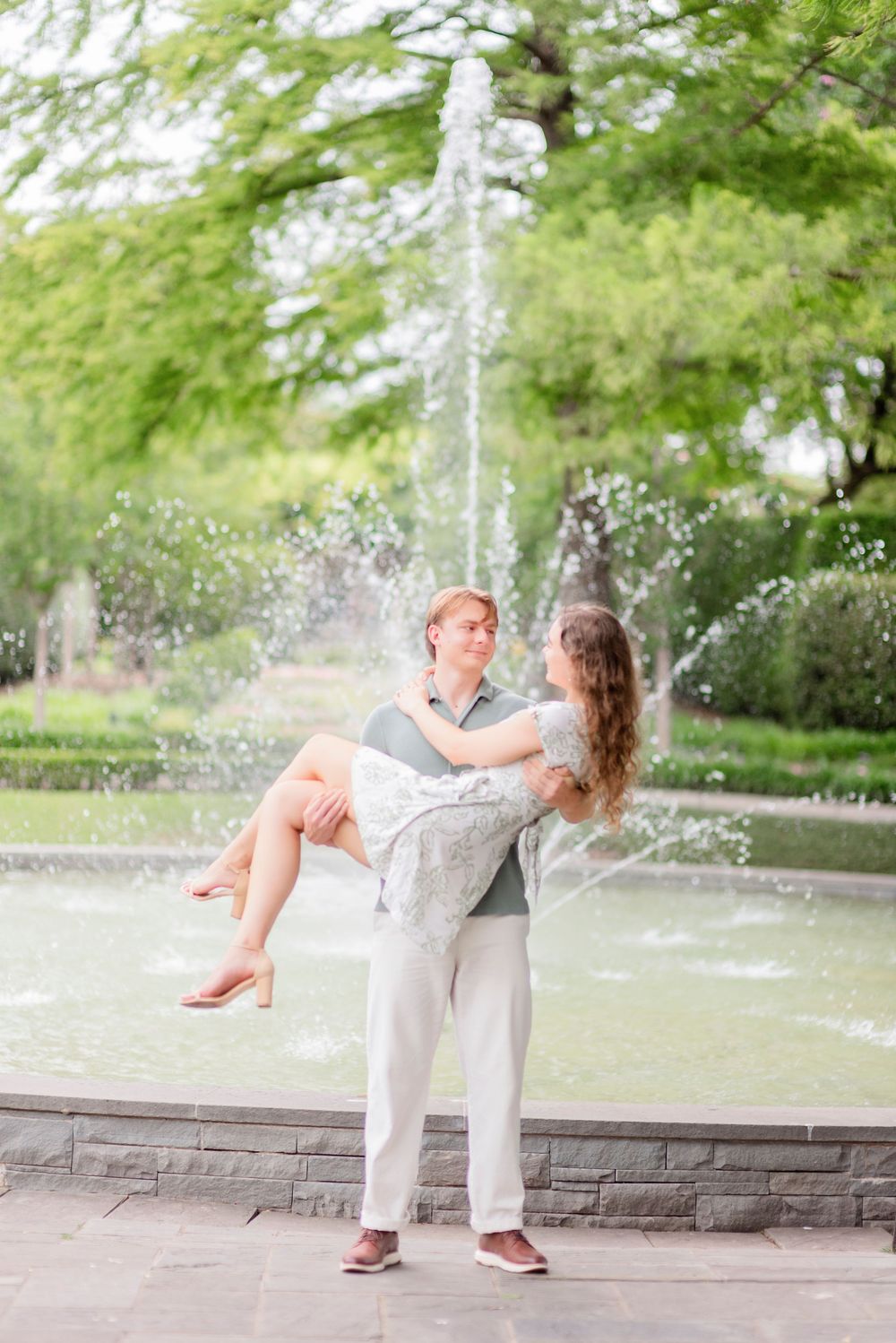 A couple shares a playful moment by a fountain as water sprays in the background of a beautiful garden.