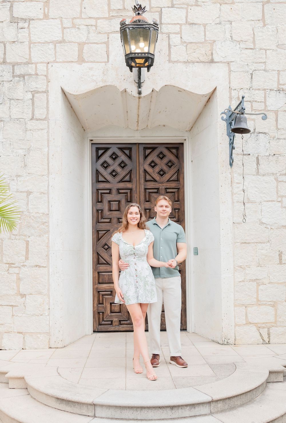 A couple poses in front of an ornate wooden door with geometric patterns and a vintage lantern overhead.