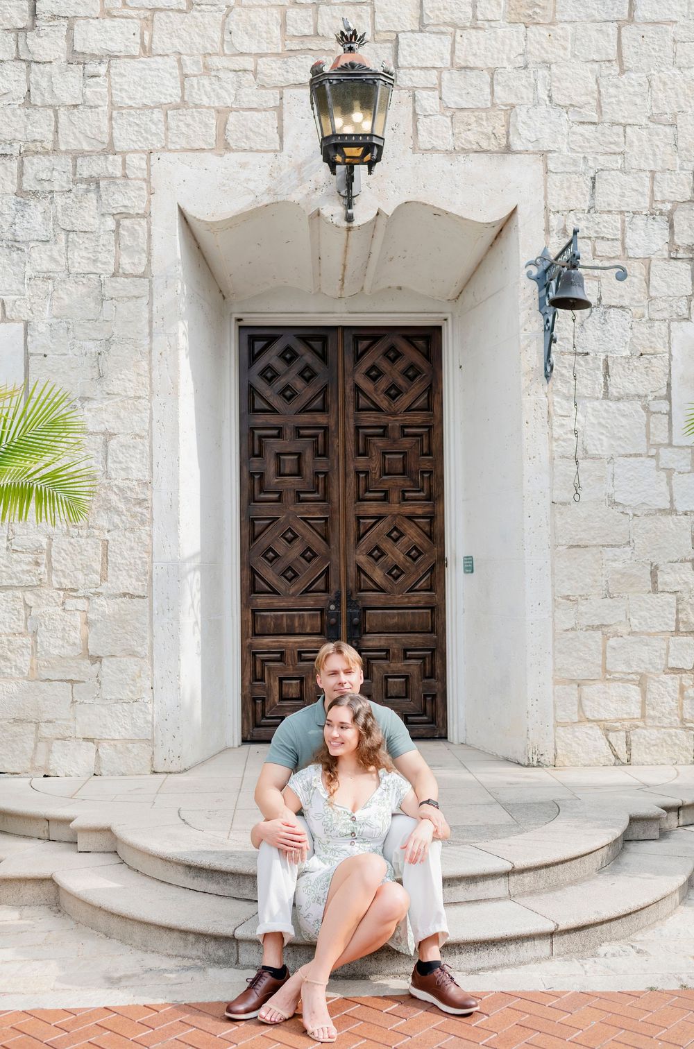 A couple sits on white stone steps in front of ornate wooden doors during a photo session.