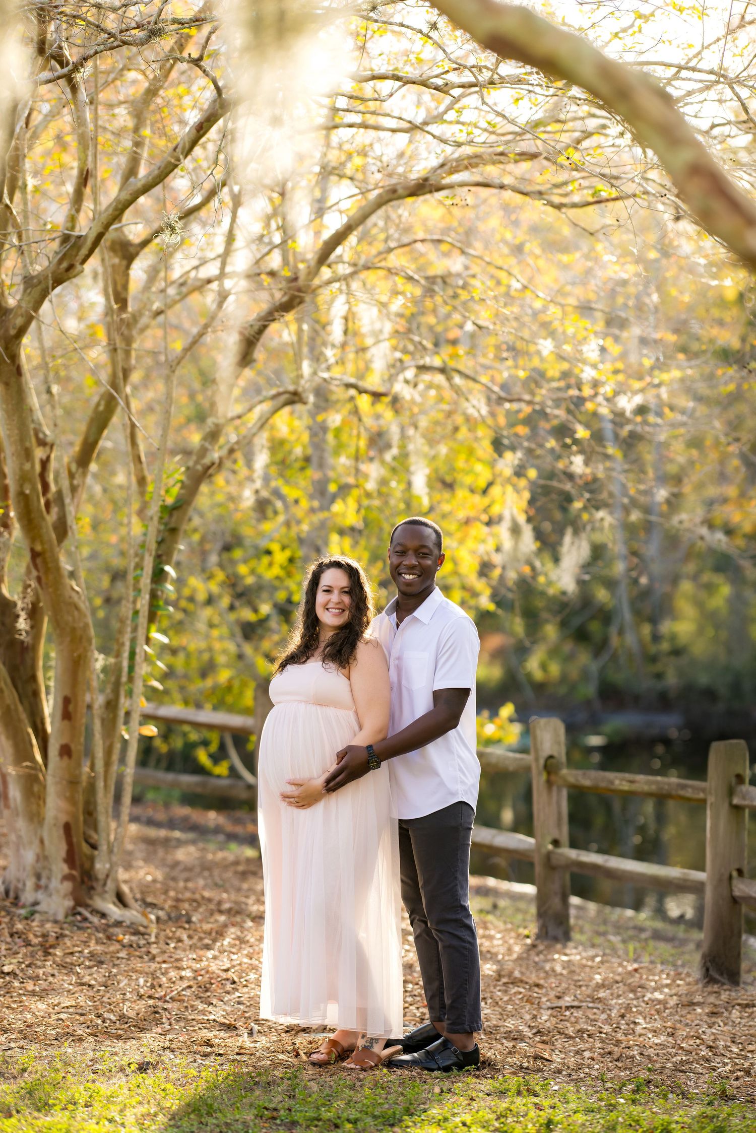 A couple embraces in a sunlit autumn forest setting with golden leaves and a rustic wooden fence.