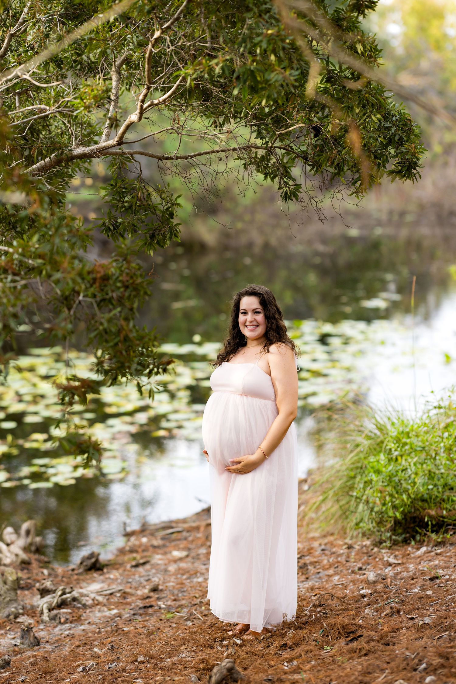 A person in a flowing white dress poses gracefully by a serene pond with lily pads and overhanging trees.