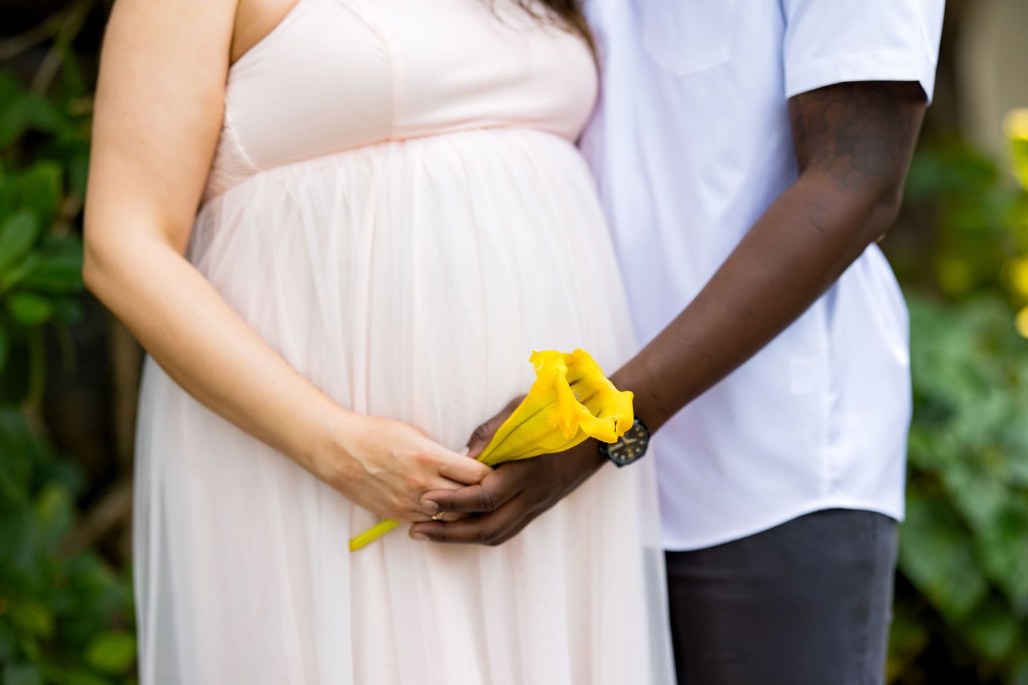A couple holds hands with yellow flowers while standing together in a garden setting, wearing white clothing.