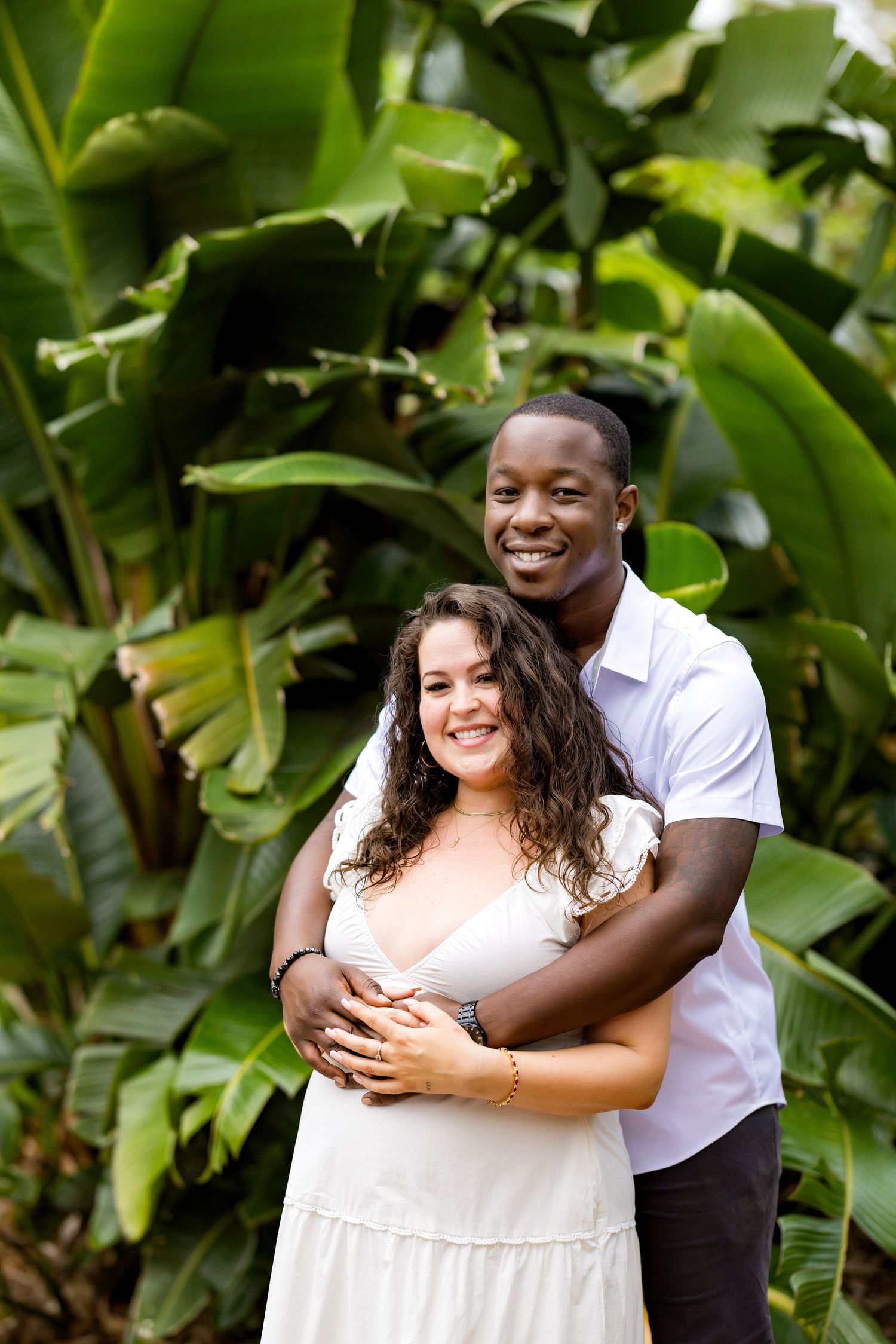 A couple embraces and smiles together in a lush tropical garden setting with green foliage in the background.