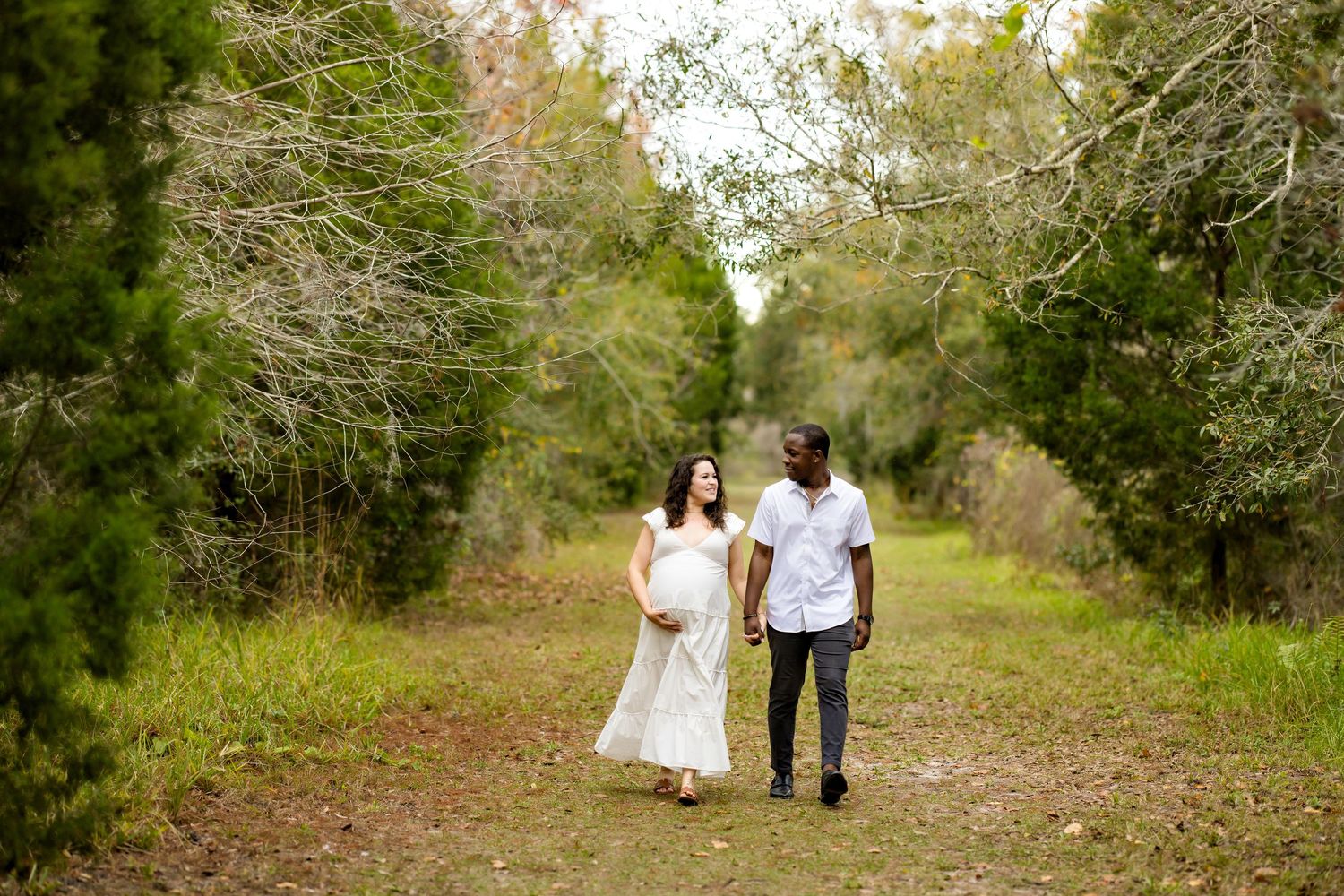 A couple walks hand in hand down a tree-lined path on a sunny day, with the woman wearing a flowing white dress.