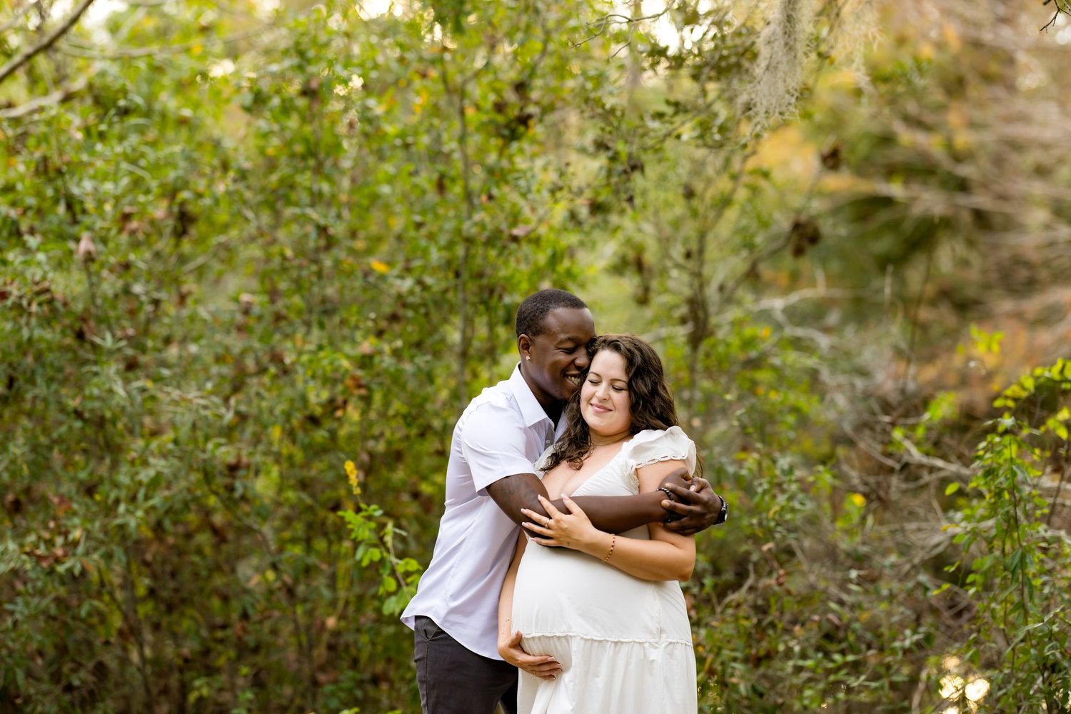 Couple embraces during outdoor photo session surrounded by lush green foliage in natural lighting.