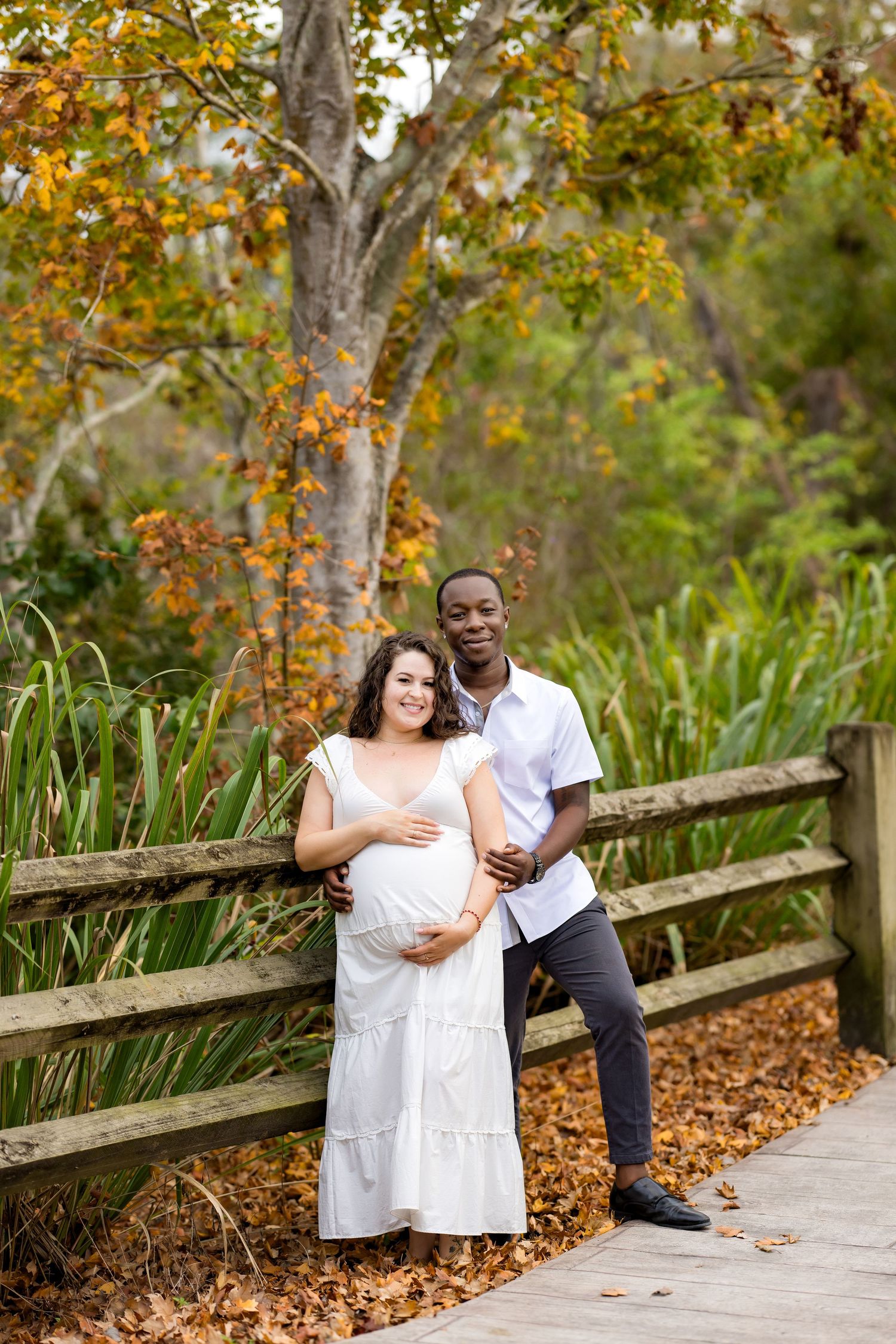 A couple stands together on a wooden walkway surrounded by autumn foliage and tall grasses in a natural setting.