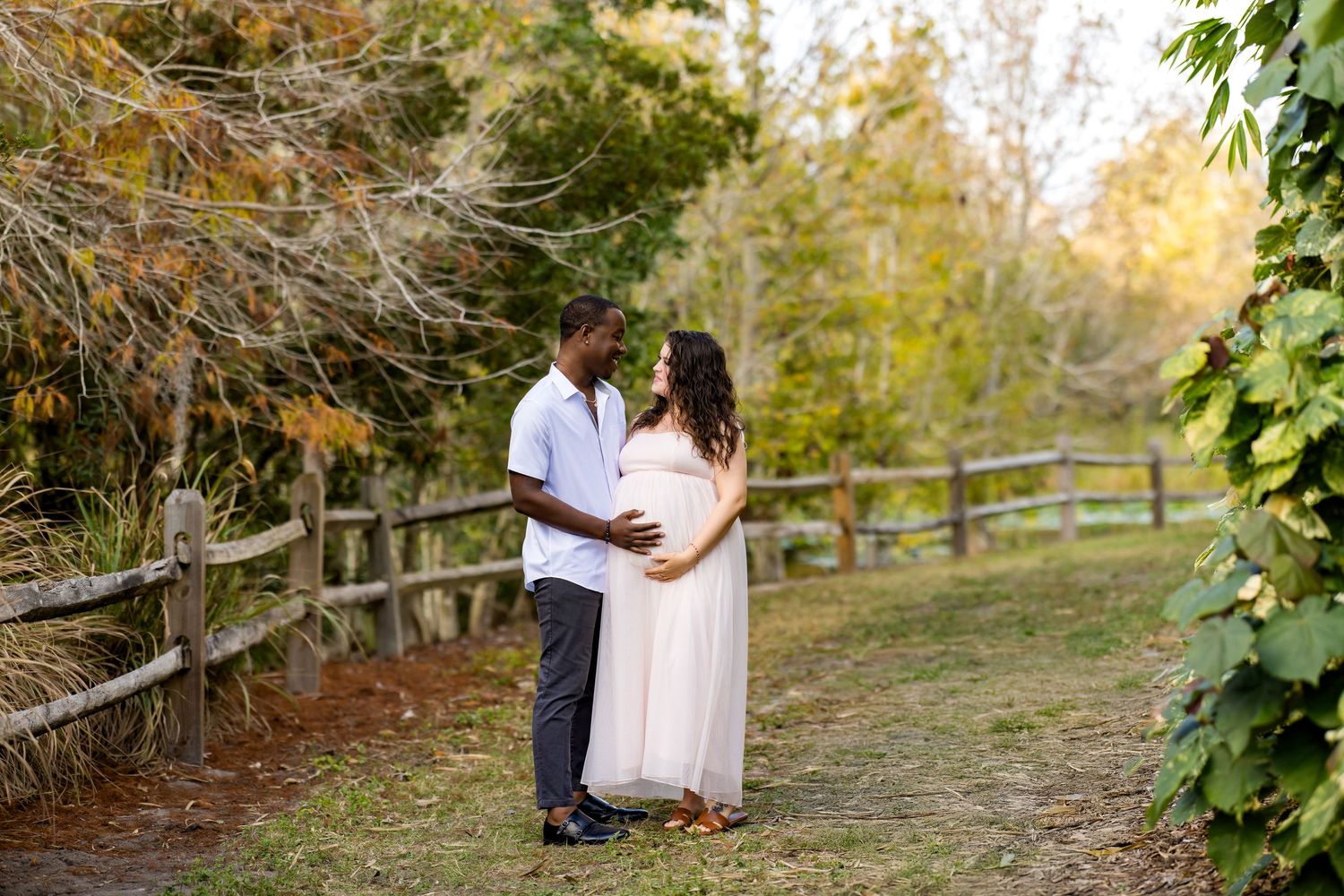 A romantic outdoor photoshoot at sunset shows a couple embracing on a rustic wooden fence in a natural setting.