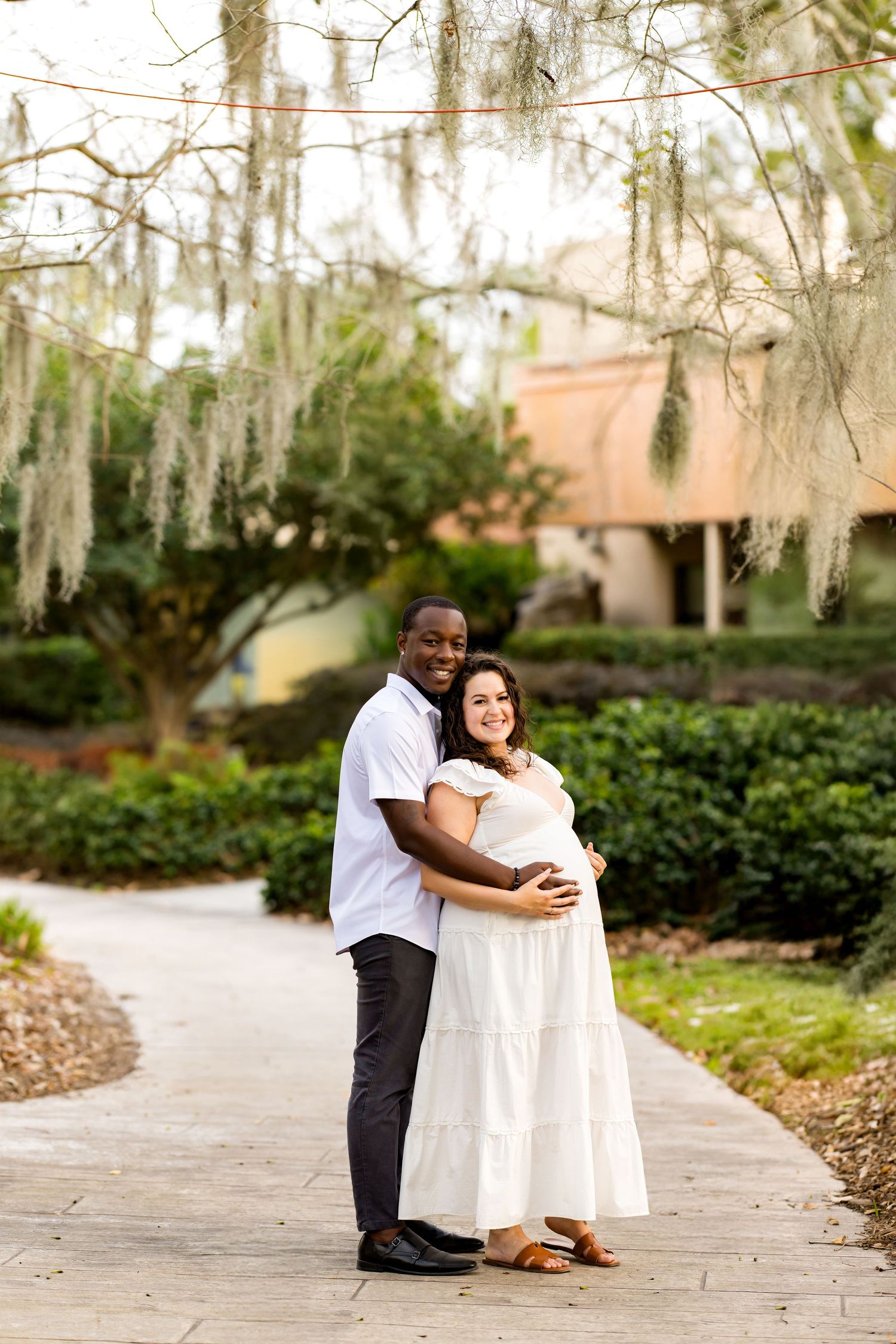 A couple shares an intimate moment together on a stone path surrounded by Spanish moss and lush gardens.