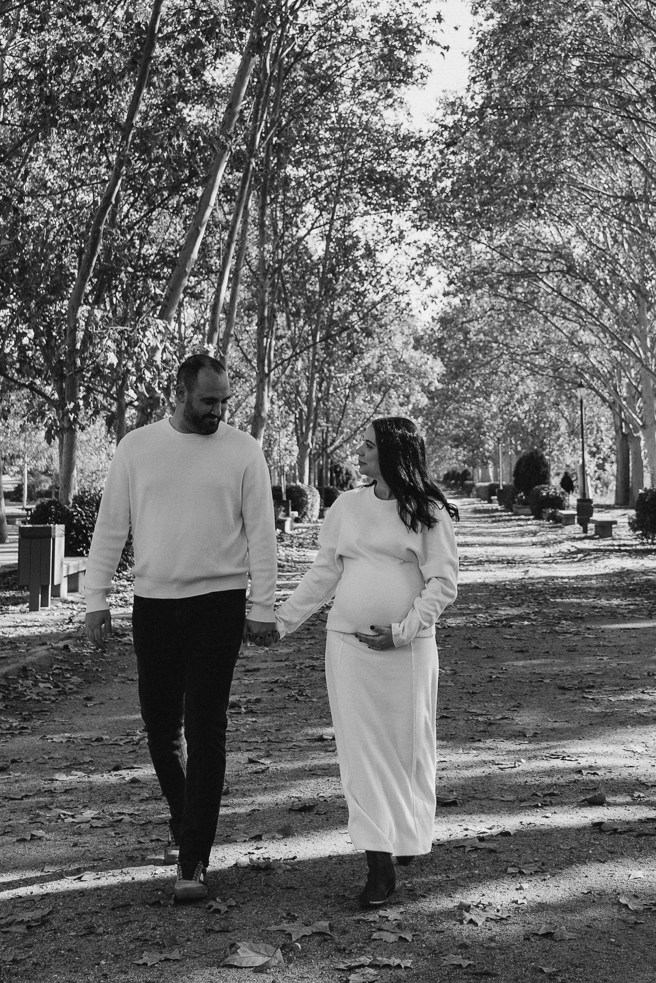 A couple walks together on a tree-lined path during autumn, with the woman wearing a flowing white dress.