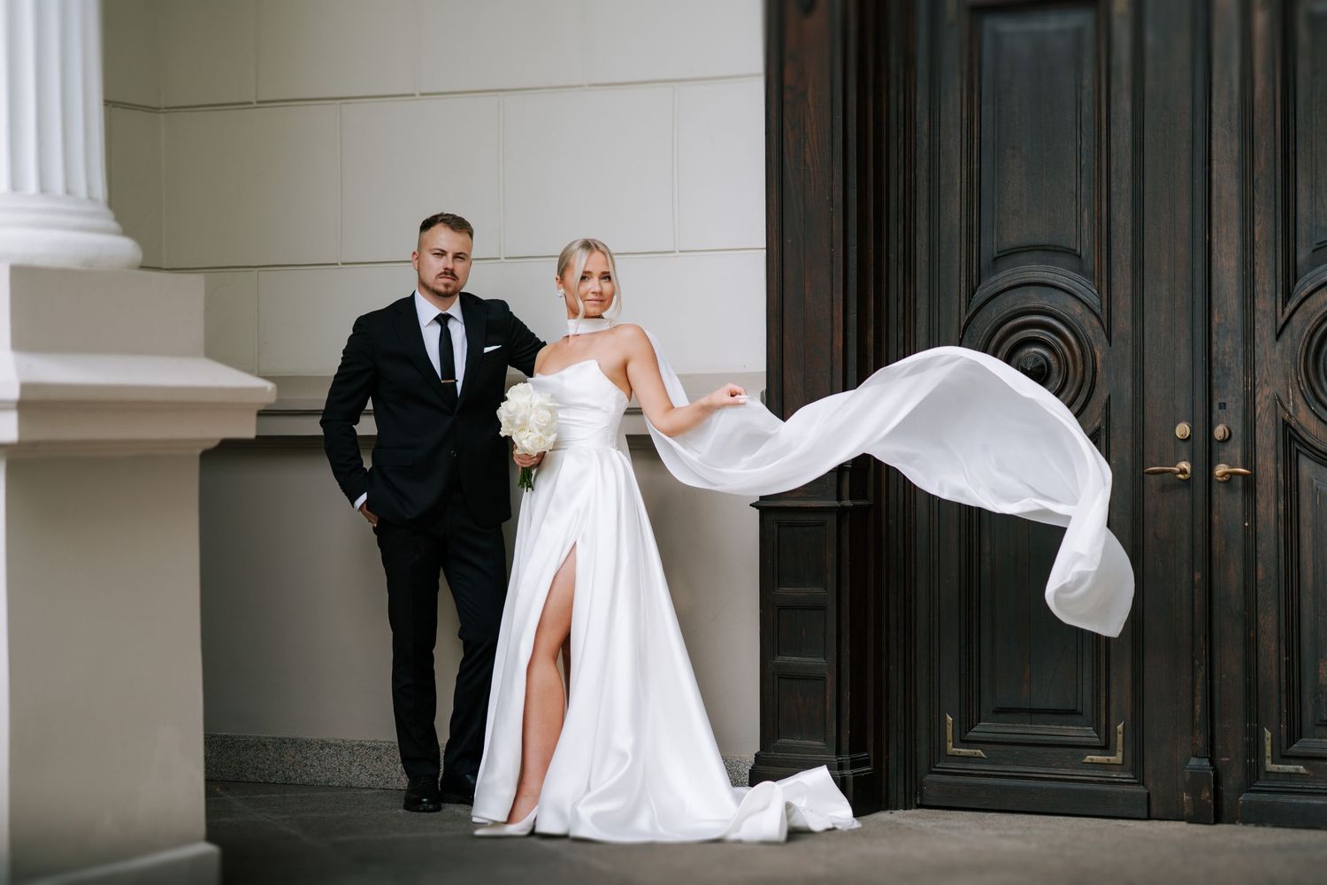 Bride's white dress flows dramatically in breeze against classic architectural backdrop.