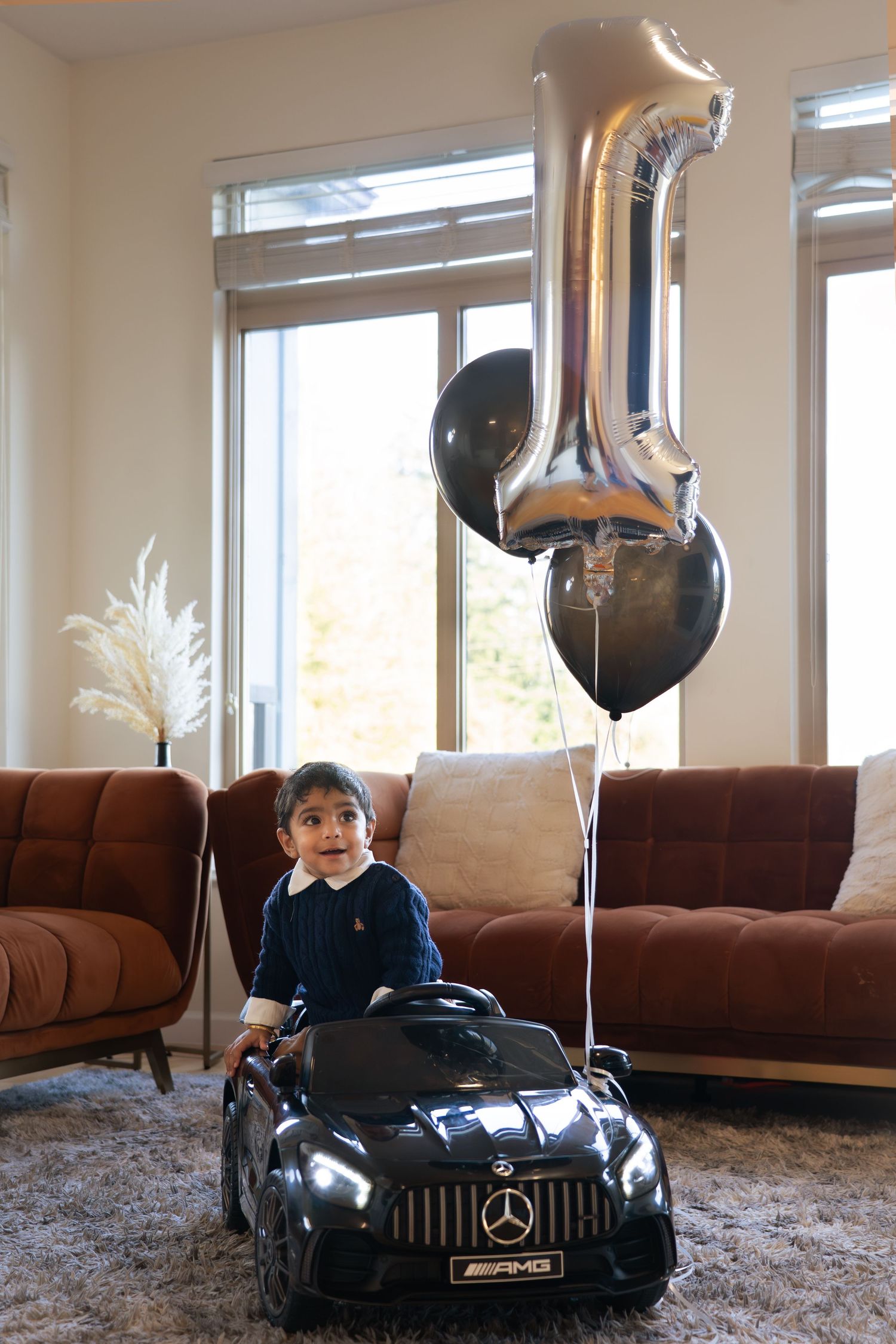 A child rides in a black toy Mercedes-Benz car on a carpeted floor with balloons in the background.