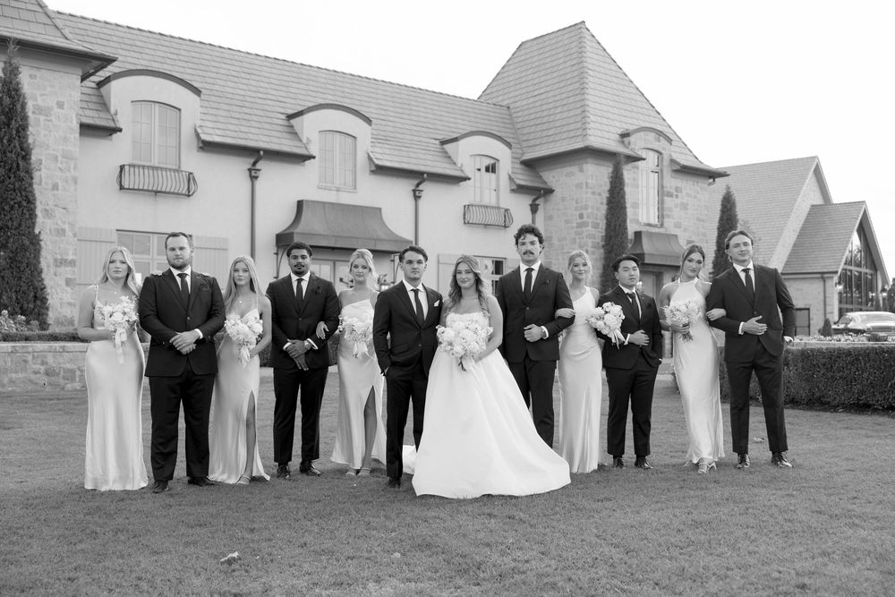 Wedding party members lined up in formal attire against a white building exterior for a group photo.