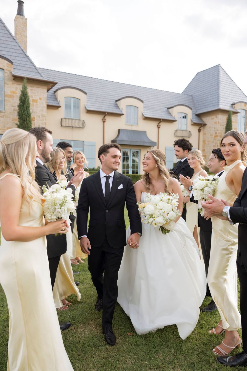 Wedding party members in formal attire walk together on grass with elegant stone building in background.