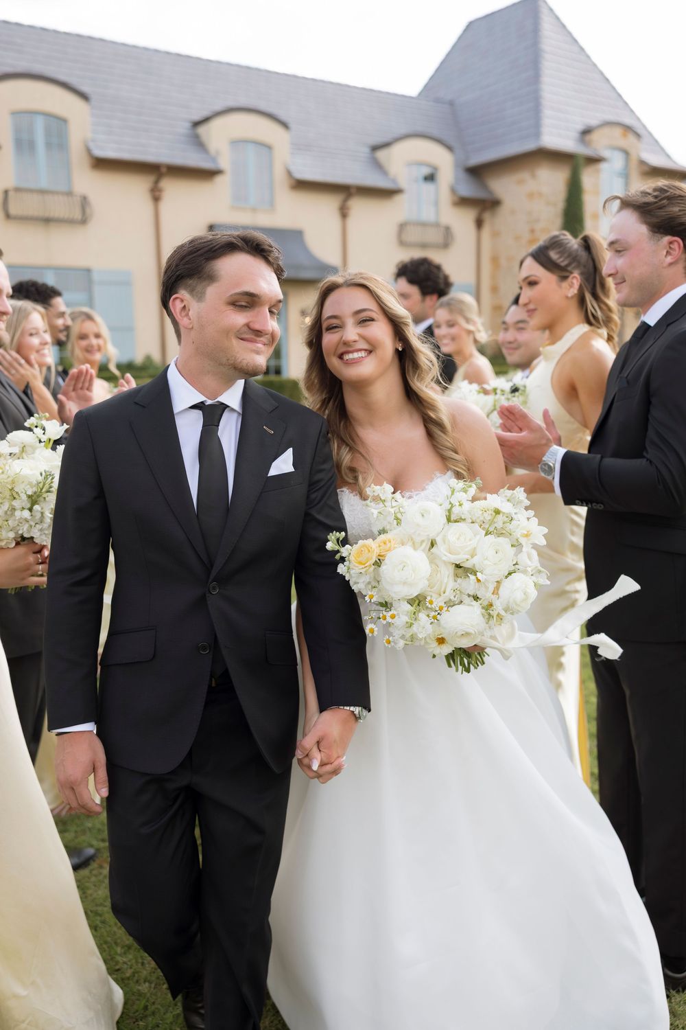 A joyful wedding celebration outdoors with a couple walking together in formal attire against a French-style mansion backdrop.