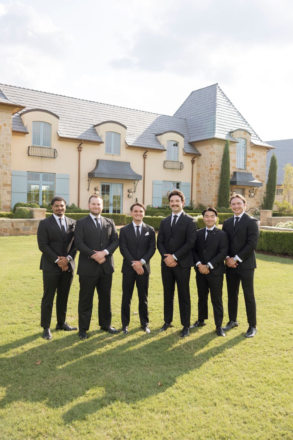 Groomsmen in black suits stand in a row on a sunny lawn casting long shadows beside a stone building.