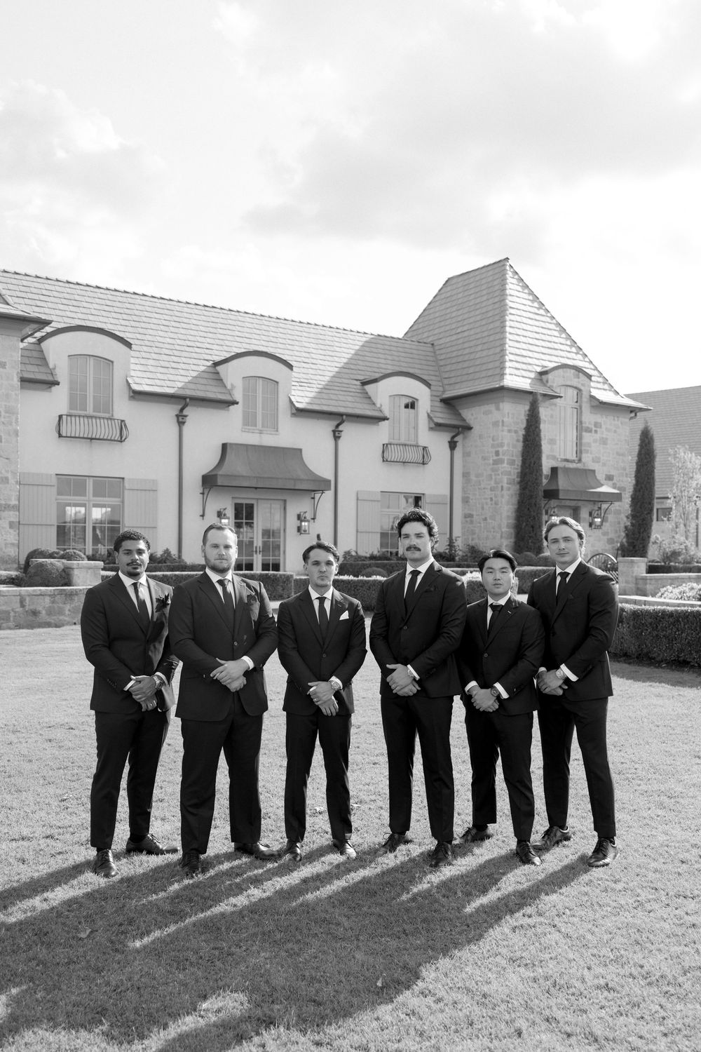 Wedding party in black suits stands in line on grass with stone building backdrop in black and white photo.
