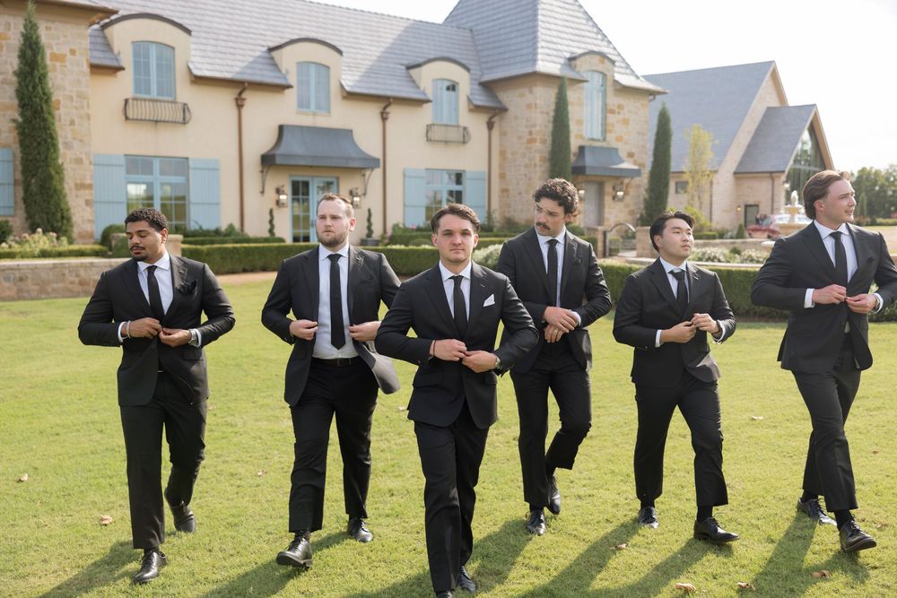 Groomsmen in dark suits perform synchronized dance moves on lawn during wedding celebration.