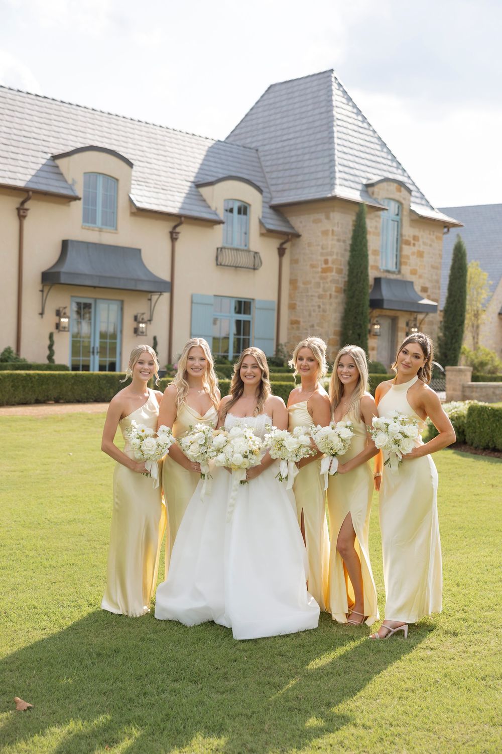 Wedding party in yellow dresses poses on sunny lawn outside elegant stone venue holding white bouquets.