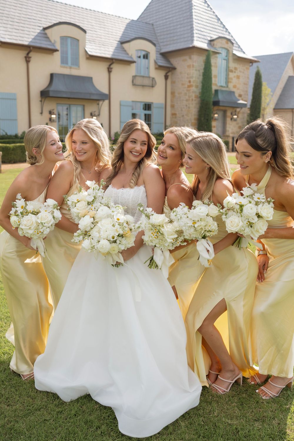 Bridesmaids in pale yellow dresses and bride in white gather on lawn holding white bouquets.