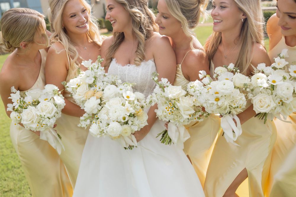 Bridesmaids in pale yellow dresses hold white floral bouquets while laughing together outdoors at a wedding celebration.