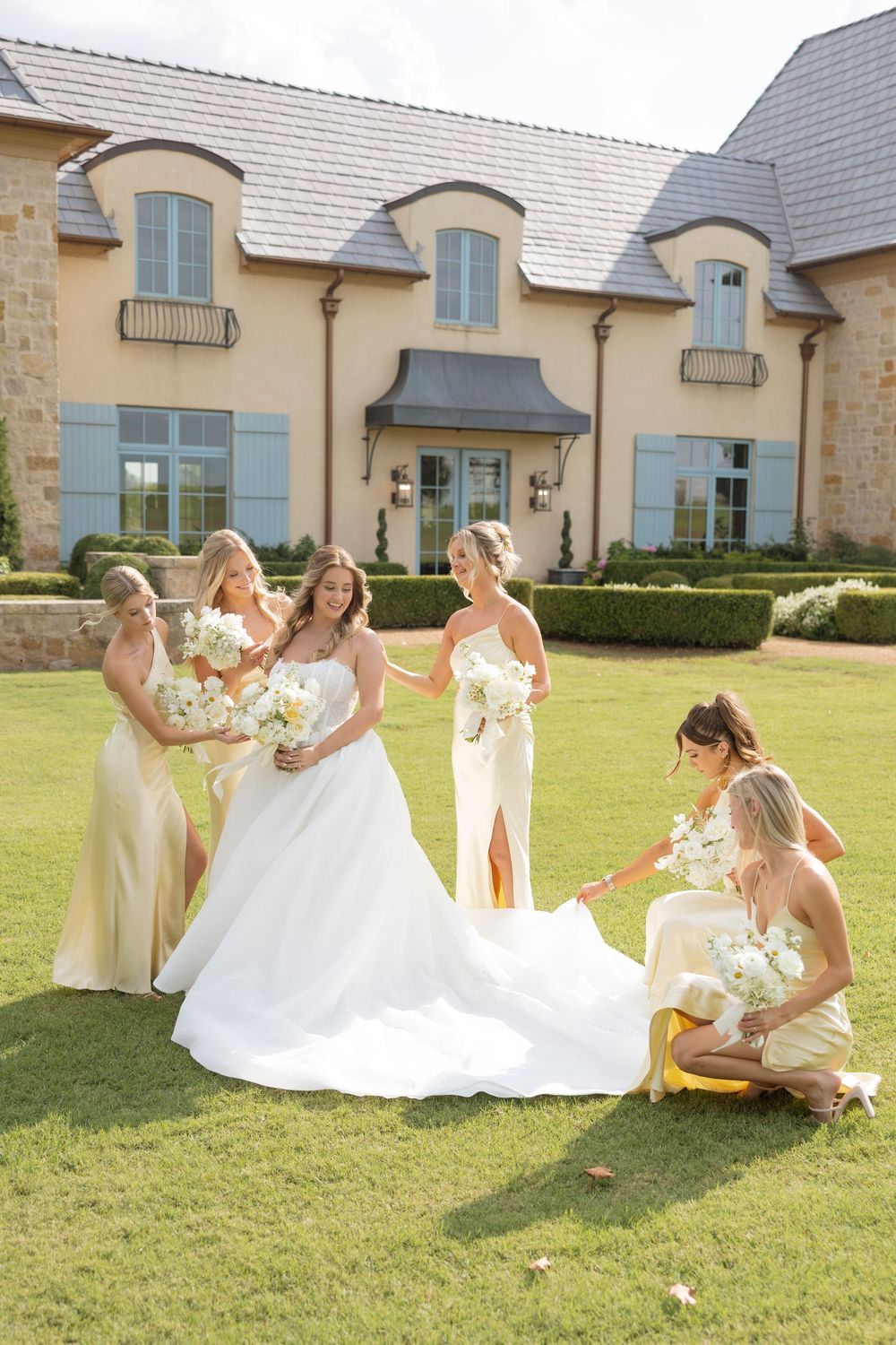 A bridal party in yellow dresses helps arrange a wedding dress on a sunny lawn outside an elegant stone building.