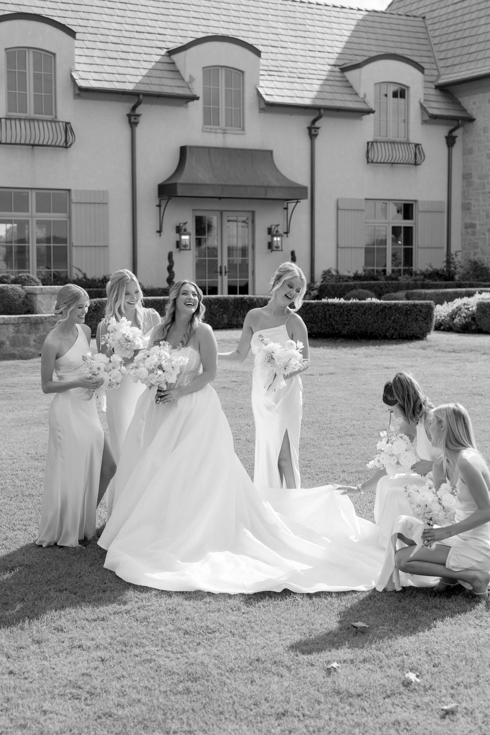 Black and white photo of bridesmaids helping arrange the bride's wedding dress train on grass.