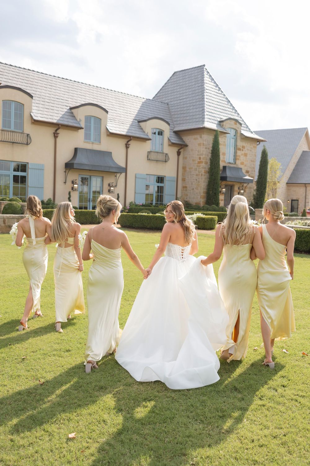 Wedding party in yellow dresses and white gown walk together across sunny lawn by French country style building.