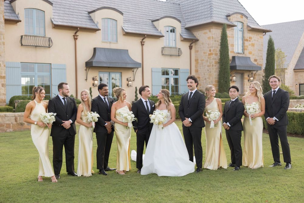 Wedding party in formal attire poses together on green lawn outside elegant stone venue during outdoor celebration.