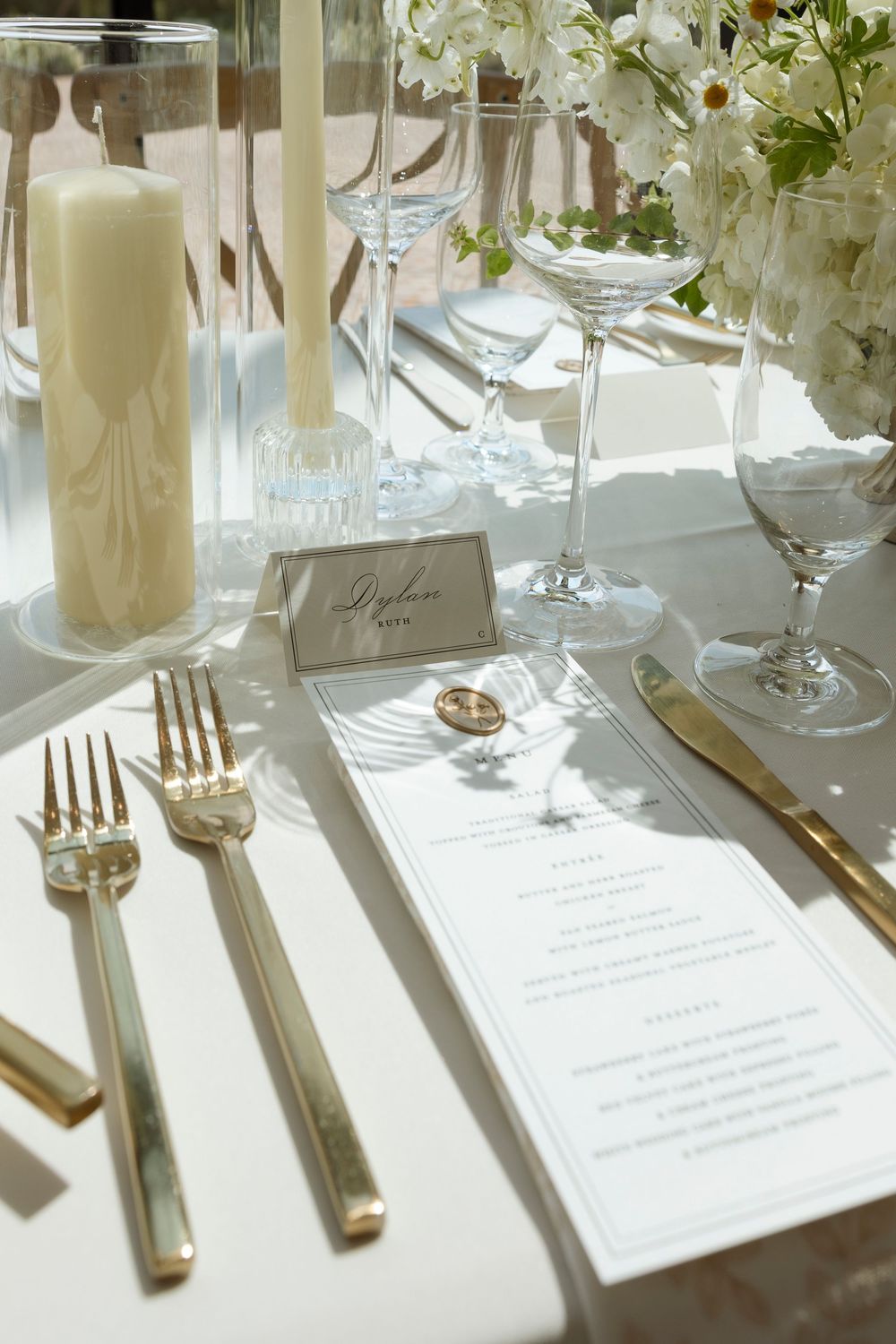 Elegant wedding place setting with white candle, menu card, gold flatware and wine glasses on white tablecloth.
