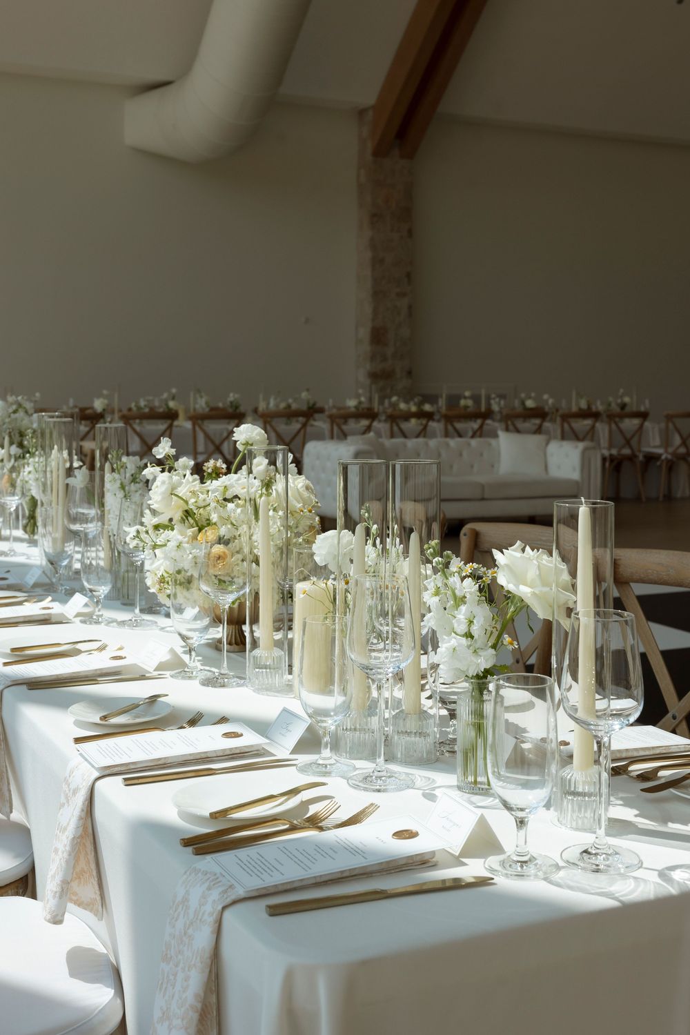 Elegant wedding reception table setting with white linens, glassware, and tall floral centerpieces in a bright indoor venue.