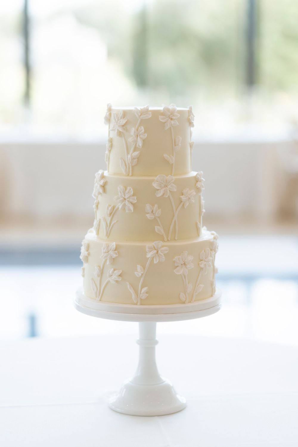 Elegant white wedding cake with delicate floral vine details displayed on a classic white pedestal stand.