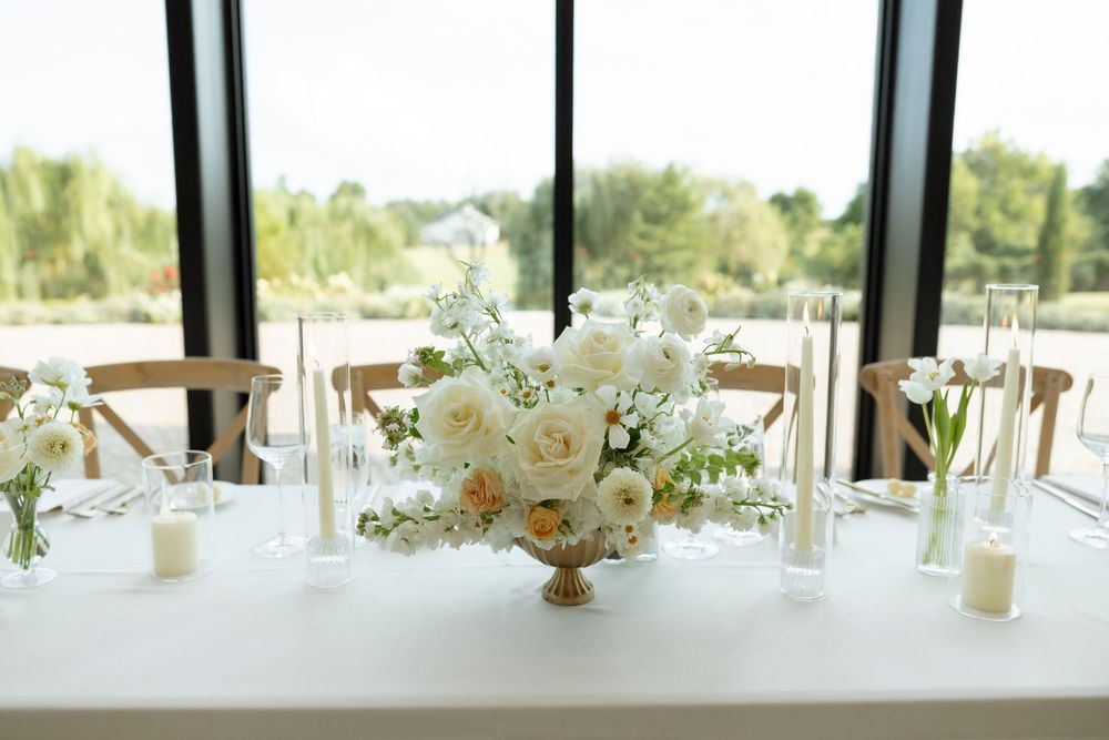 Long banquet tables with white floral centerpieces and cross-back chairs in a bright reception hall with panoramic windows.