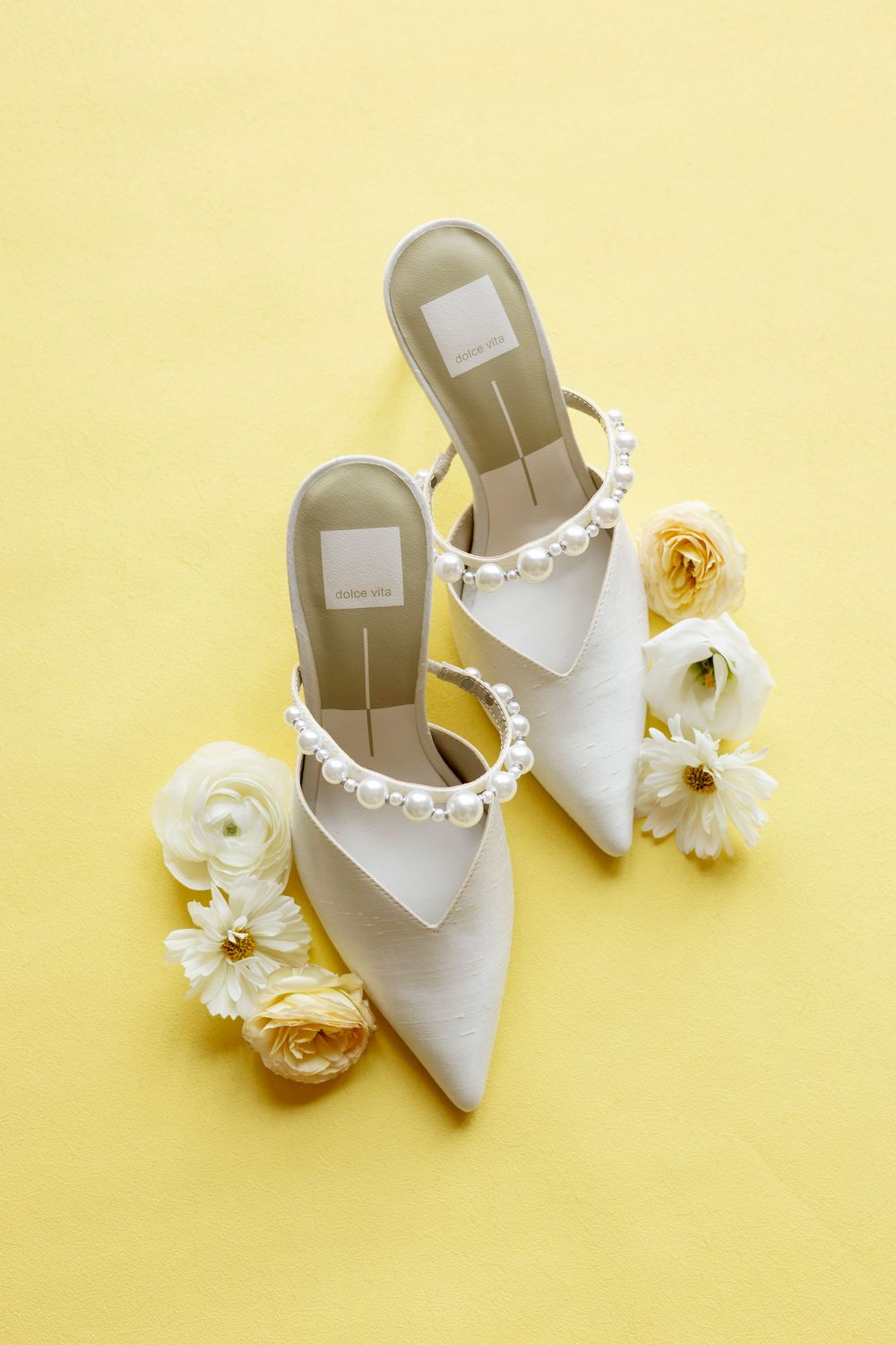 White bridal heels with delicate embellishments paired with cream-colored roses against a soft yellow background.