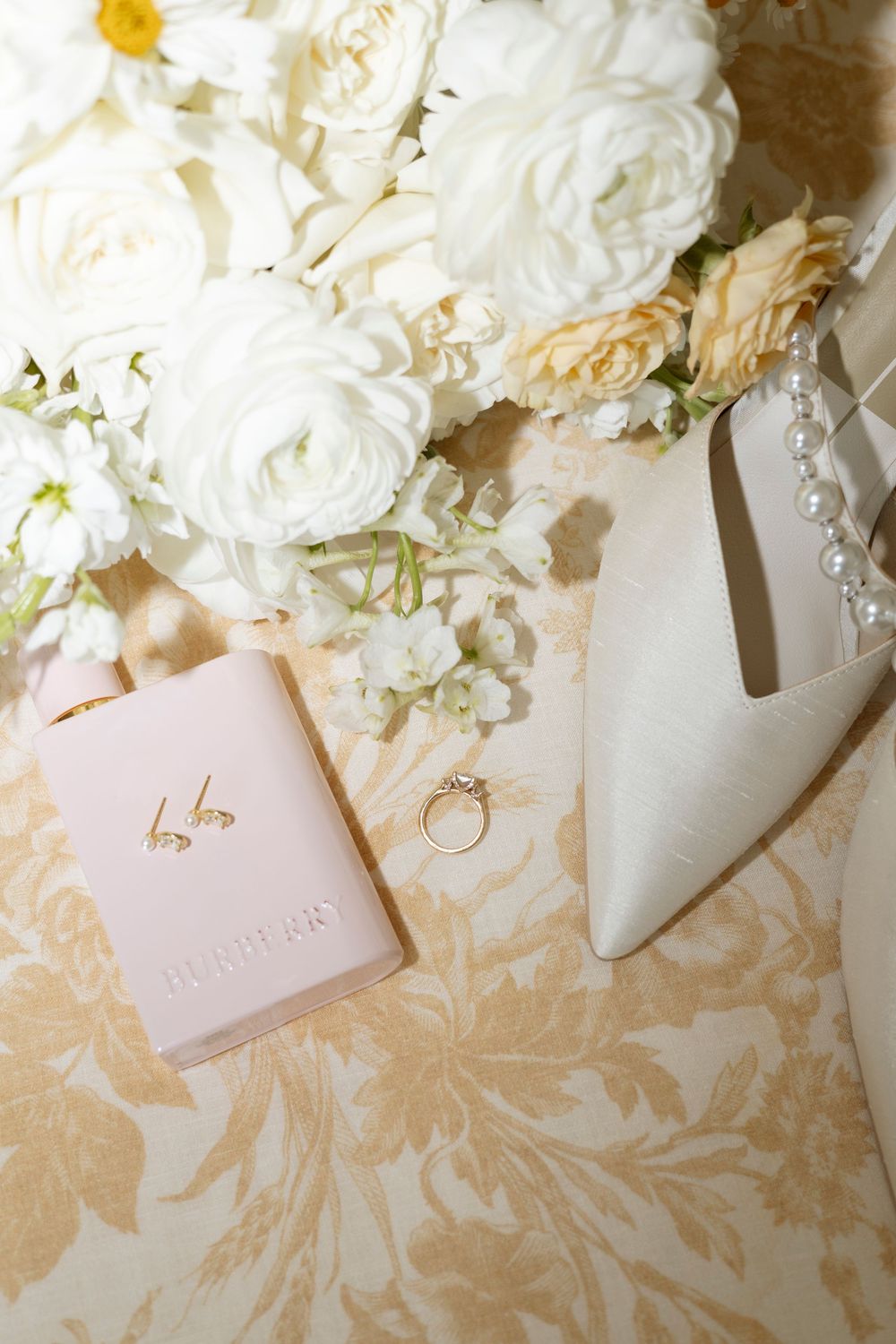 Wedding details flatlay with white roses, engagement ring, and bridal shoes on a beige floral background.