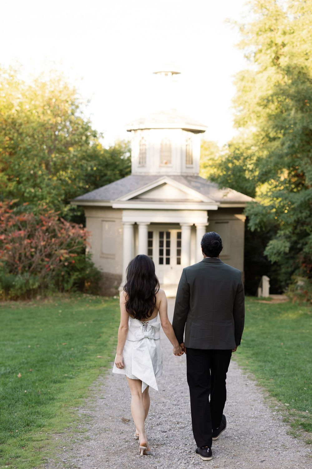 A couple in formal attire walking away on a garden path towards a white building on a sunny day.