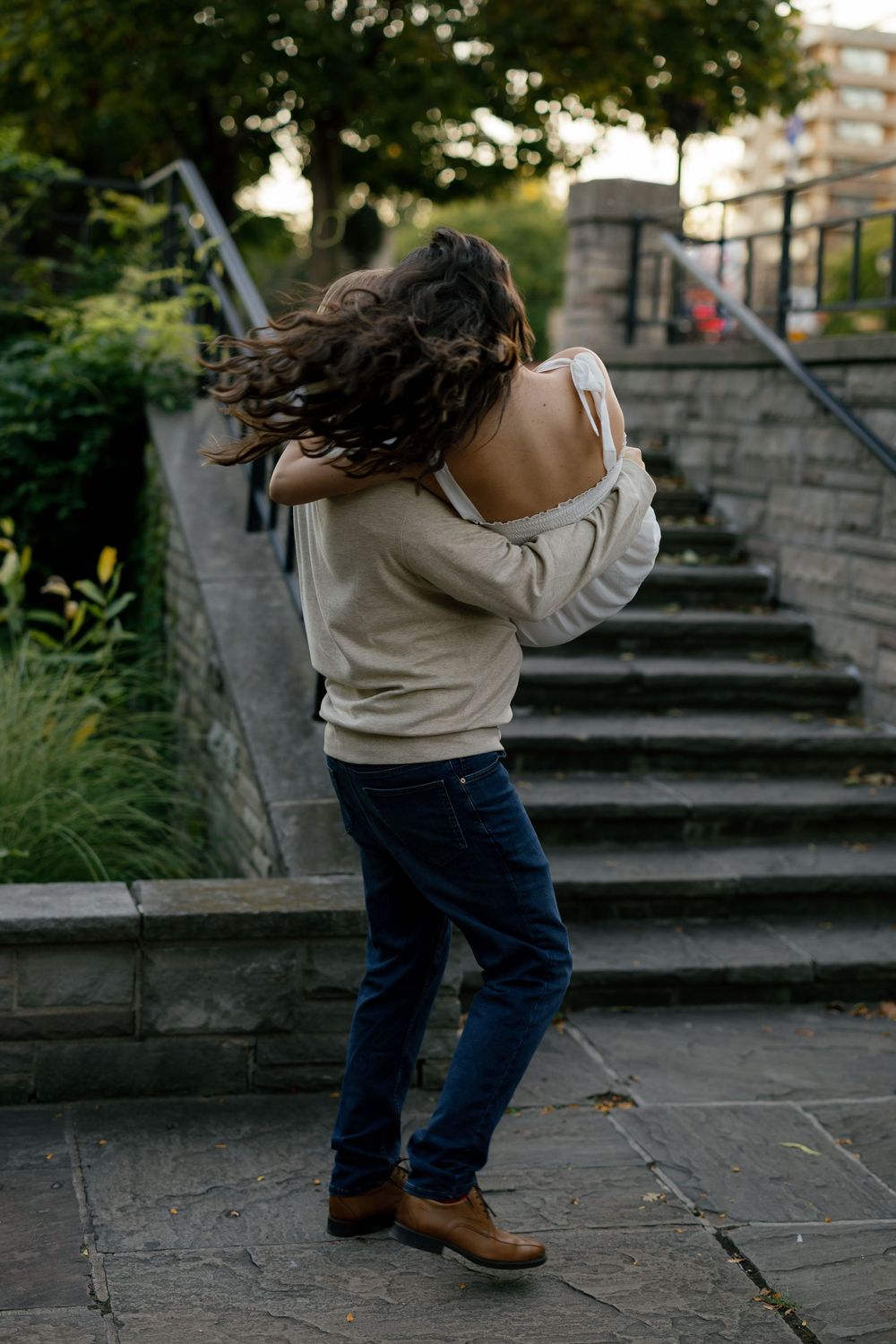 A person in jeans and a beige sweater spins around playfully on outdoor stone steps in an urban setting.